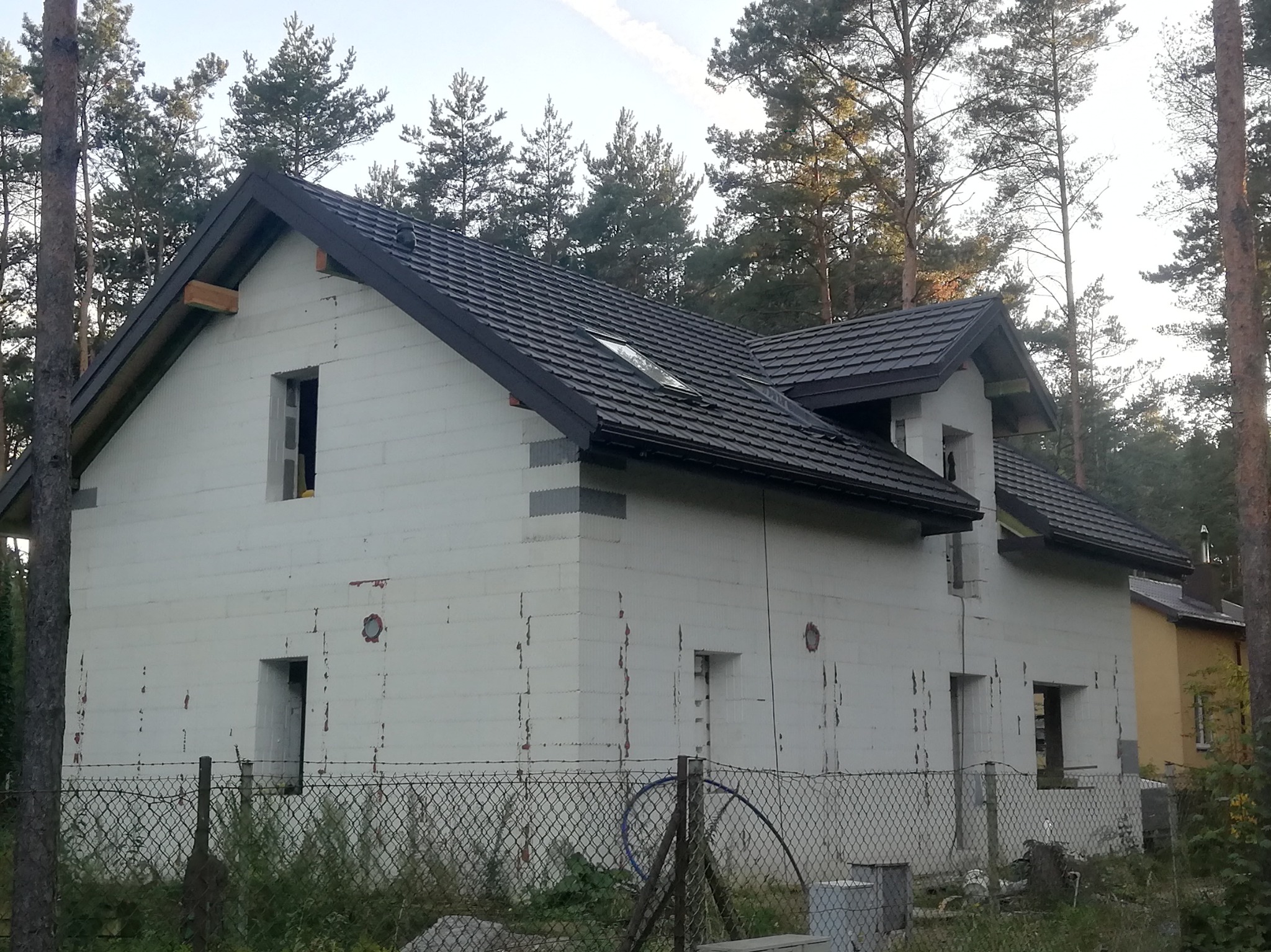 Dom w stanie surowym zamkniętym z ciemną dachówką, widoczne okna dachowe i niezadaszone otwory okienne, otoczony drzewami i ogrodzeniem z siatki.