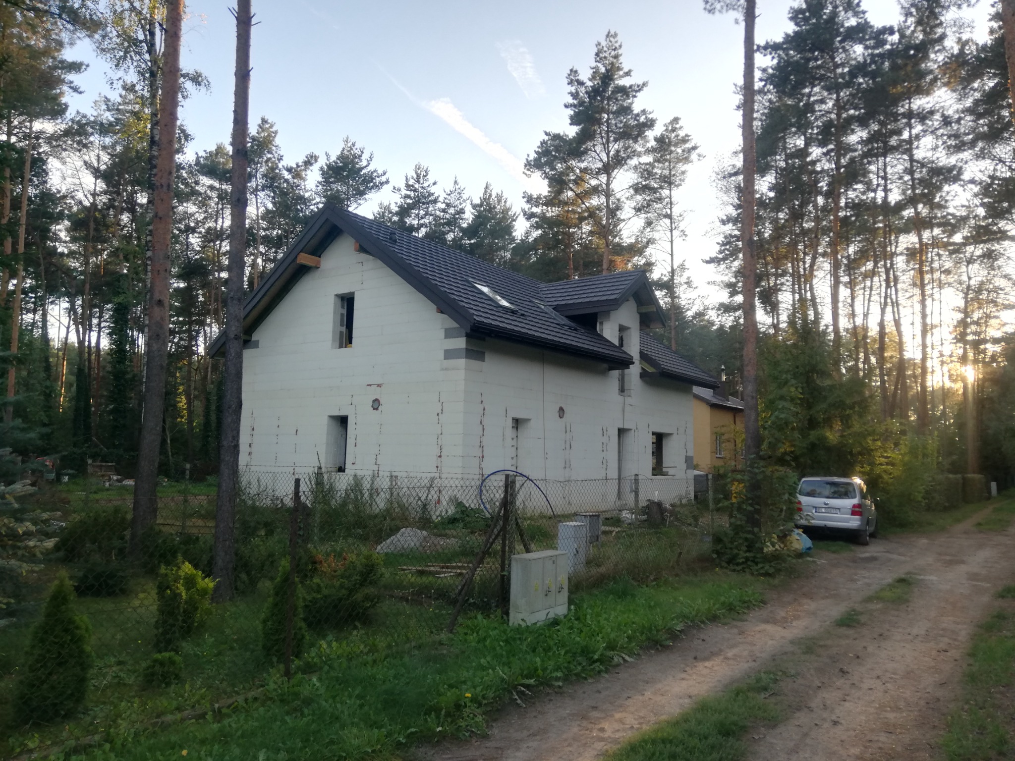 Dom w trakcie budowy, ocieplony styropianem, z zamontowaną ciemną dachówką, otoczony lasem i ogrodzeniem z siatki.