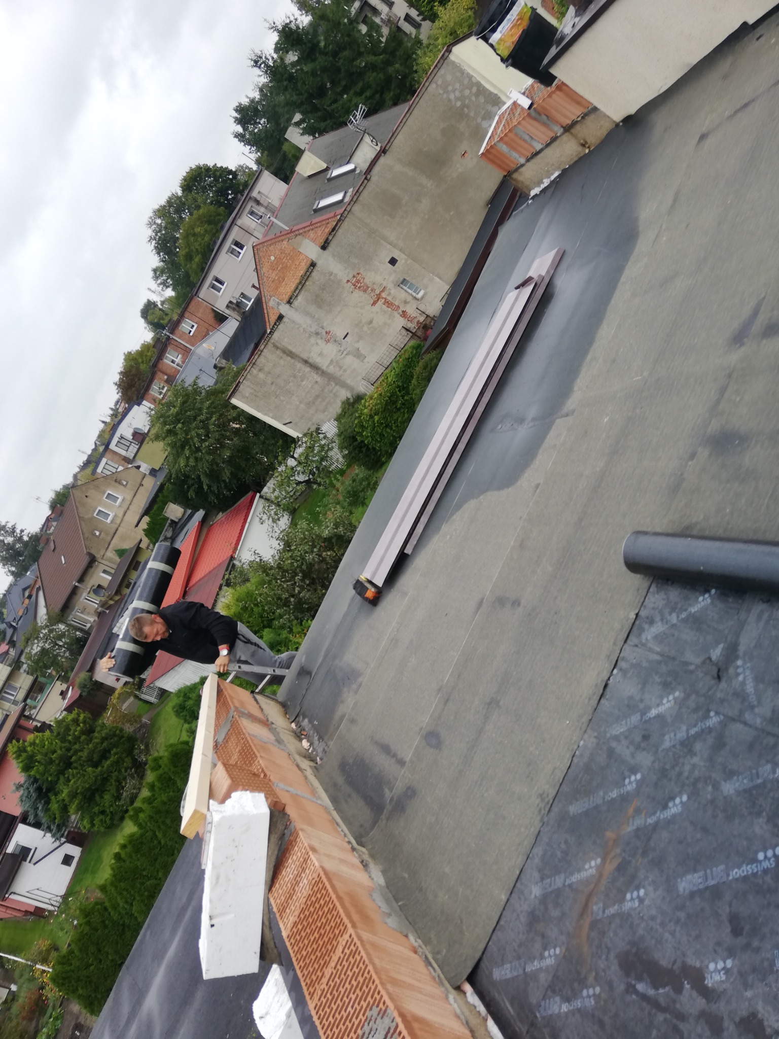 Pracownik na dachu podczas montażu obróbek blacharskich, widoczne elementy konstrukcyjne i uszczelniające, w tle zabudowa miejska.