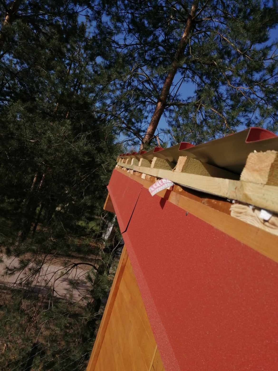 Fragment czerwonego dachu z widoczną izolacją z wełny mineralnej, drewnianą konstrukcją więźby i blacharskimi obróbkami, na tle drzew iglastych i błękitnego nieba.