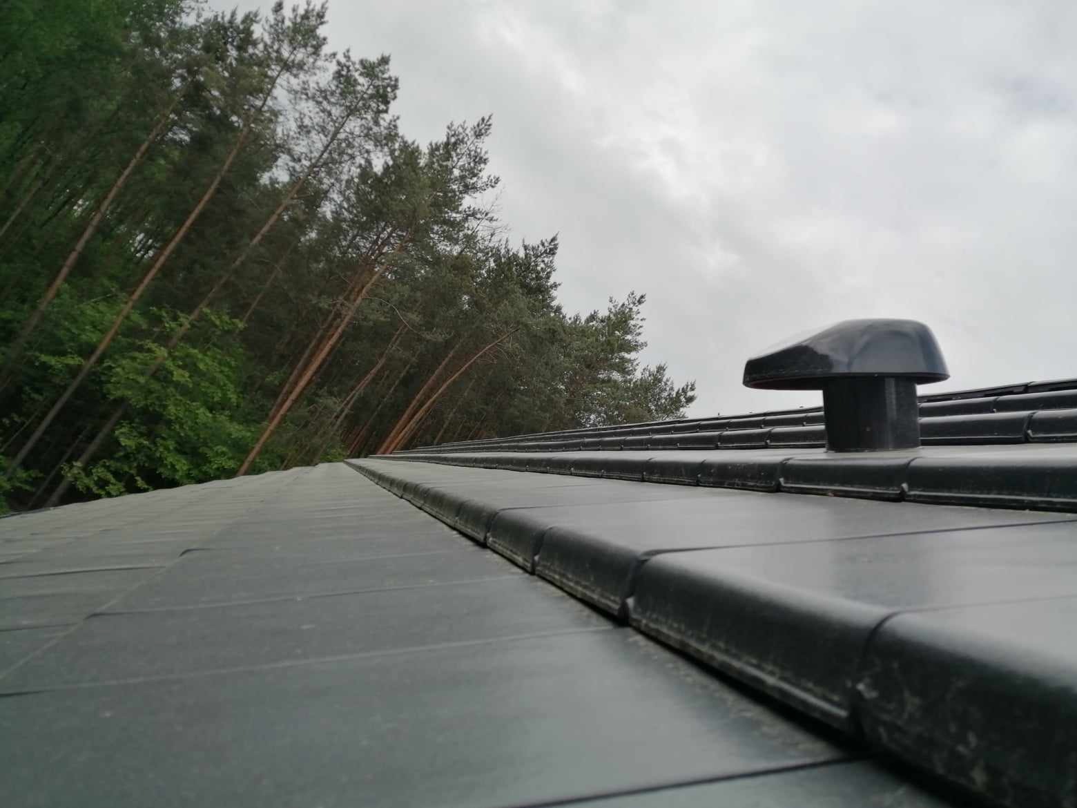 Nowoczesny, grafitowy dach z ceramicznej dachówki z zamontowanym odpowietrznikiem na tle lasu iglastego i pochmurnego nieba.