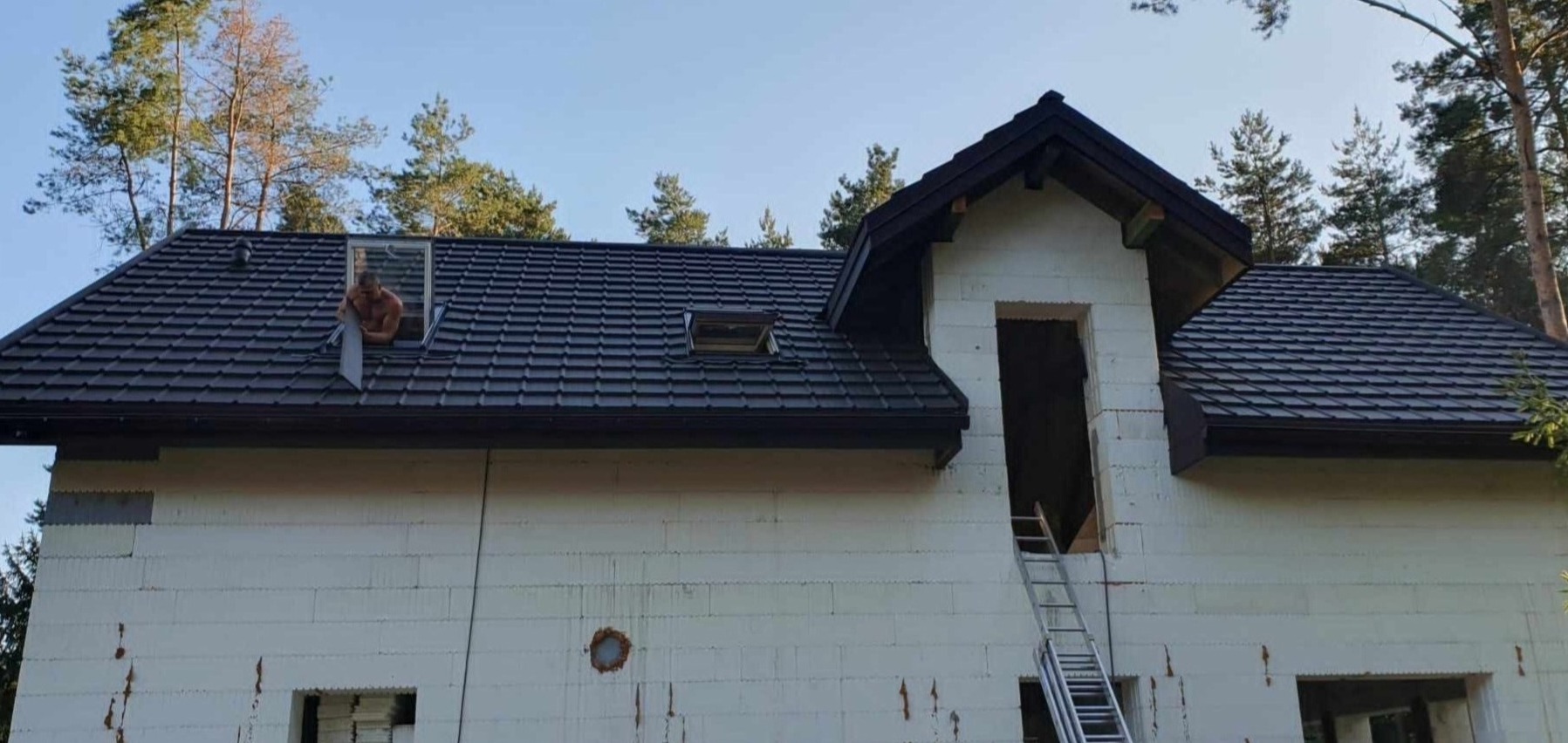 Montaż okna dachowego w nowym, ciemnoszarym dachu z blachodachówki na budynku w trakcie budowy; widoczny dekarz w otworze okiennym.