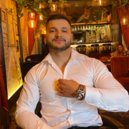 Mężczyzna w białej koszuli zapinający guzik, siedzący w eleganckim wnętrzu restauracji z dekoracyjnymi lampami i roślinami.