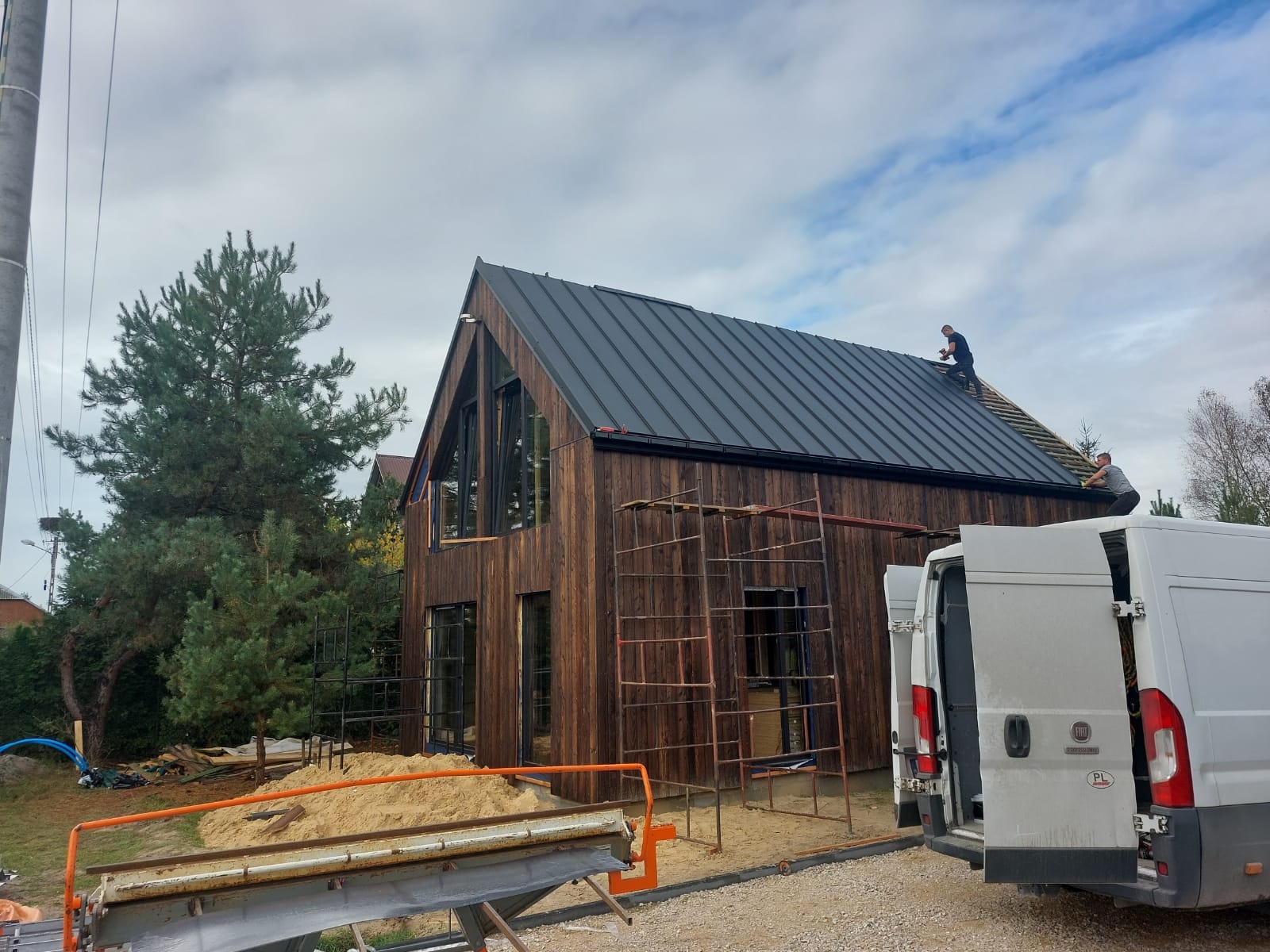Montaż blachy na dachu nowoczesnego domu z ciemnego drewna, widoczne rusztowania i samochód dostawczy z otwartymi drzwiami.