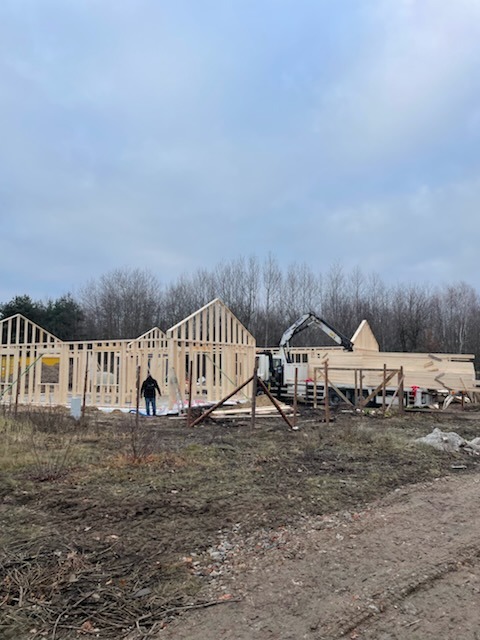 Wznoszenie drewnianej konstrukcji domu z belek, widoczny dźwig i pracownik na placu budowy.