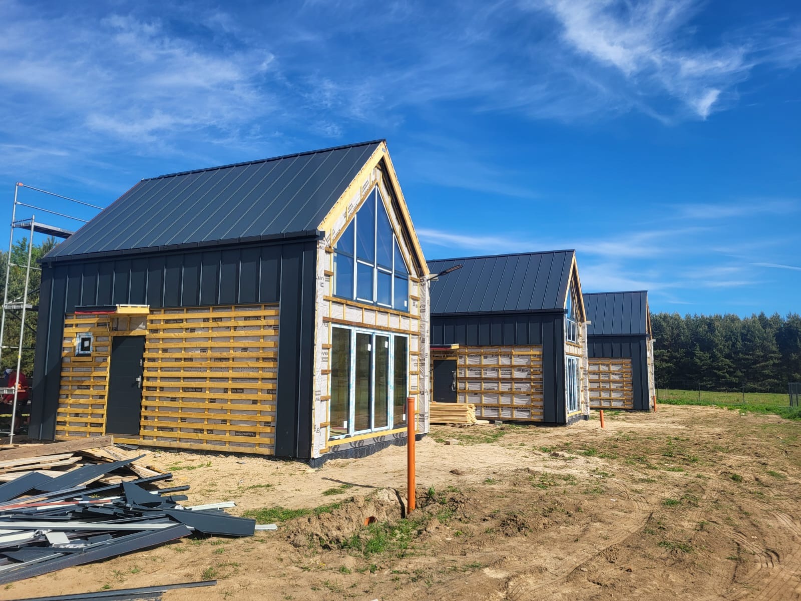 Domy szkieletowe w trakcie budowy w Wodzinie Prywatnym, widoczne trzy budynki z szarym dachem i drewnianą elewacją, częściowo zabezpieczone folią, na pierwszym planie stos blach i wykop.