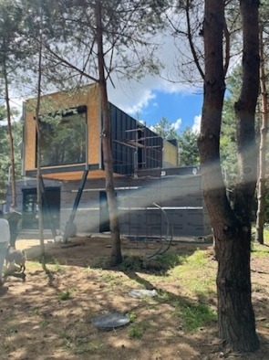 Wznoszona konstrukcja dwukondygnacyjnego budynku z elementami szkieletowymi, widoczna w lesie sosnowym, z częściowo wykończoną elewacją i stalowymi podporami.
