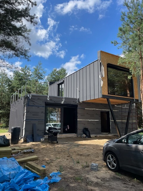 Wznoszony dwukondygnacyjny budynek w technologii szkieletowej z szarą elewacją, widoczne materiały izolacyjne, rusztowania i samochód osobowy na pierwszym planie.