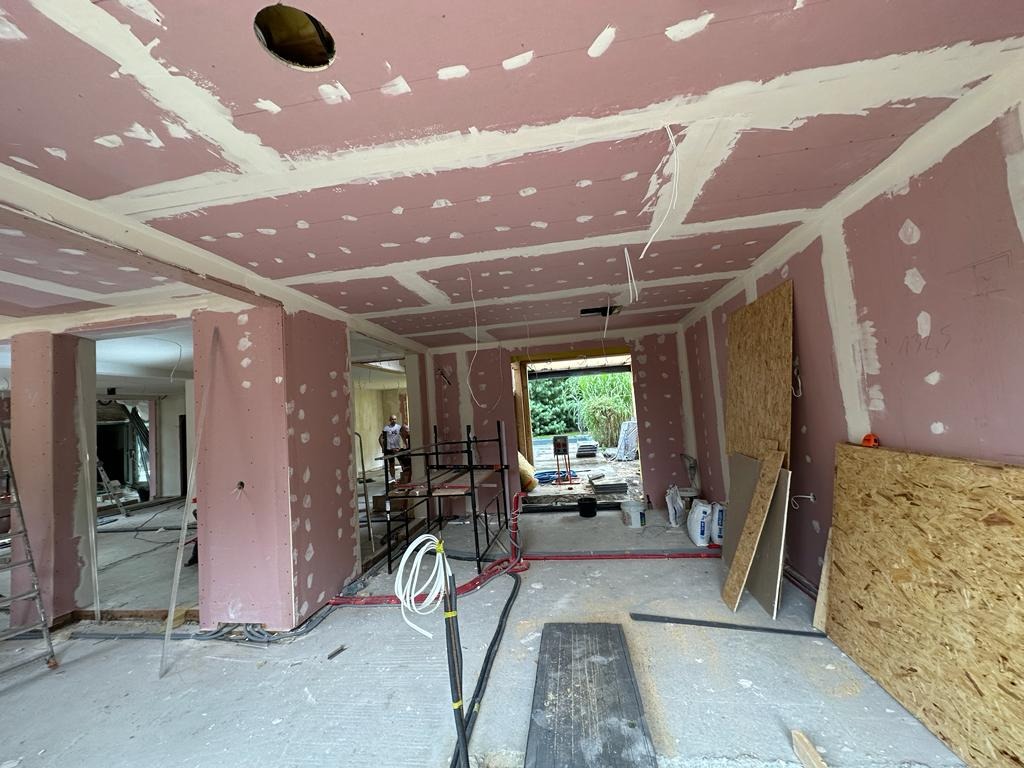 Wnętrze w trakcie wykańczania: różowe płyty gipsowo-kartonowe na ścianach i suficie, widoczne łączenia szpachlowane na biało, instalacje elektryczne, rusztowania, płyty drewniane, widok na zewnątrz...