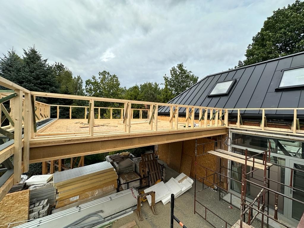 Konstrukcja drewniana łącząca budynek z nową częścią, widok z góry, metalowy profil nośny, materiały budowlane składowane na dole, czarny dach z oknami połaciowymi.