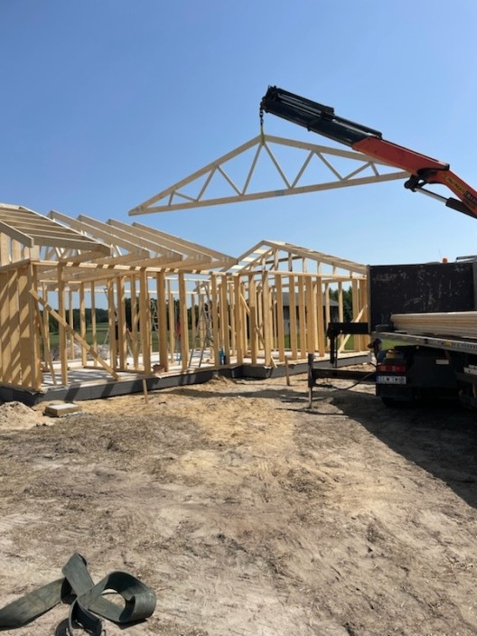Montaż drewnianej więźby dachowej za pomocą dźwigu na placu budowy, widoczne elementy konstrukcyjne ścian i fundamentów.