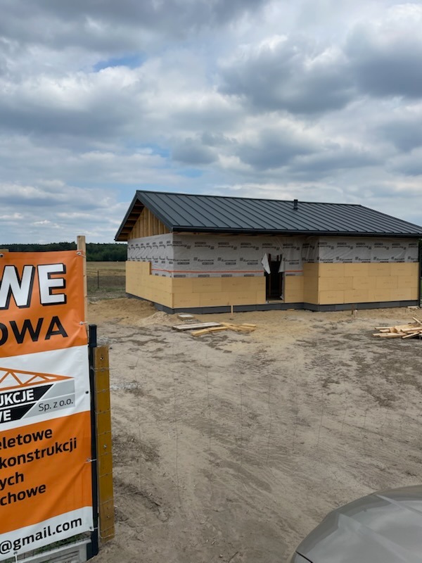 Budowa domu w technologii szkieletowej na etapie montażu ścian zewnętrznych z izolacją, widoczny czarny dach i teren budowy z tablicą reklamową firmy.