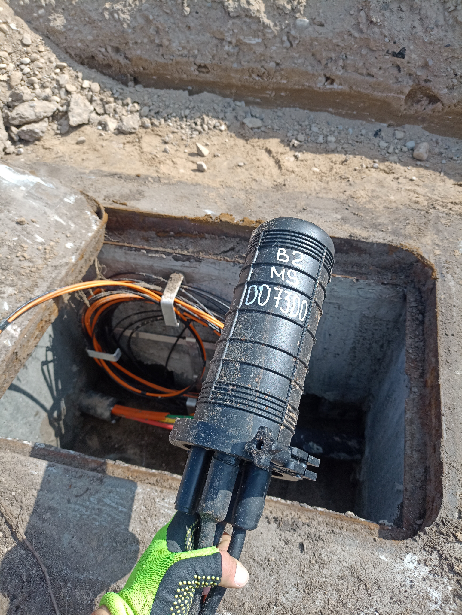 Czarna mufa światłowodowa z napisem B2 MS 007300 trzymana w rękawicy roboczej na tle betonowej studzienki z kablami światłowodowymi.