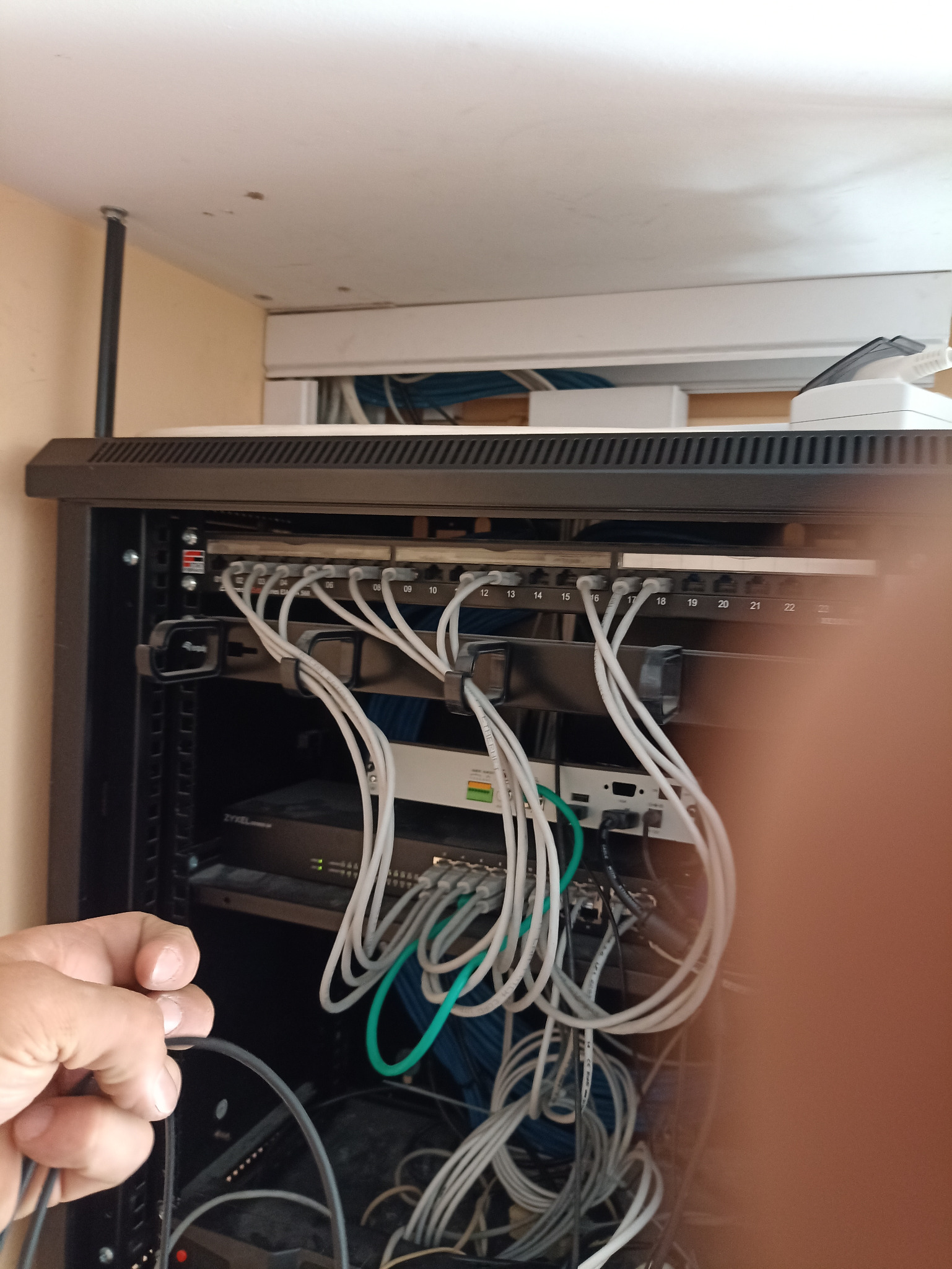 Panel krosowniczy z podłączonymi kablami sieciowymi w szafie rack, widoczne oznaczenia portów od 08 do 22, z elementami marki Zyxel, instalator trzyma w ręku kabel ethernet.