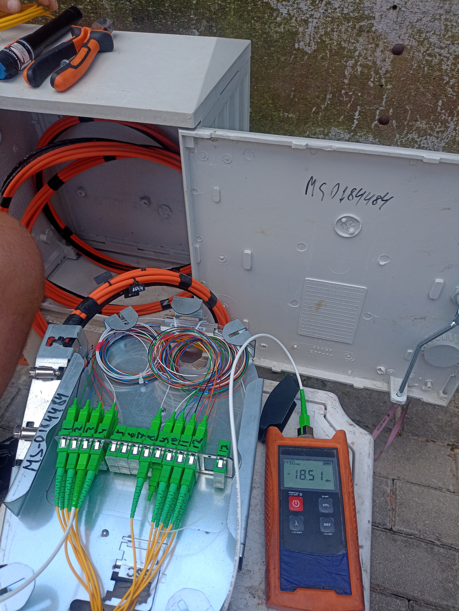 Otwarta skrzynka telekomunikacyjna z pomarańczowymi kablami i zielonymi złączami światłowodowymi, pomiar mocy sygnału za pomocą miernika optycznego, narzędzia obok.