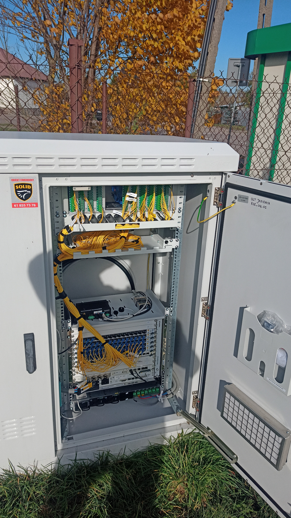 Otwarta szafa telekomunikacyjna z panelami krosowniczymi, światłowodami i urządzeniami sieciowymi, widoczny kabel uziemiający, na zewnątrz szafy trawa i drzewo z żółtymi liśćmi.
