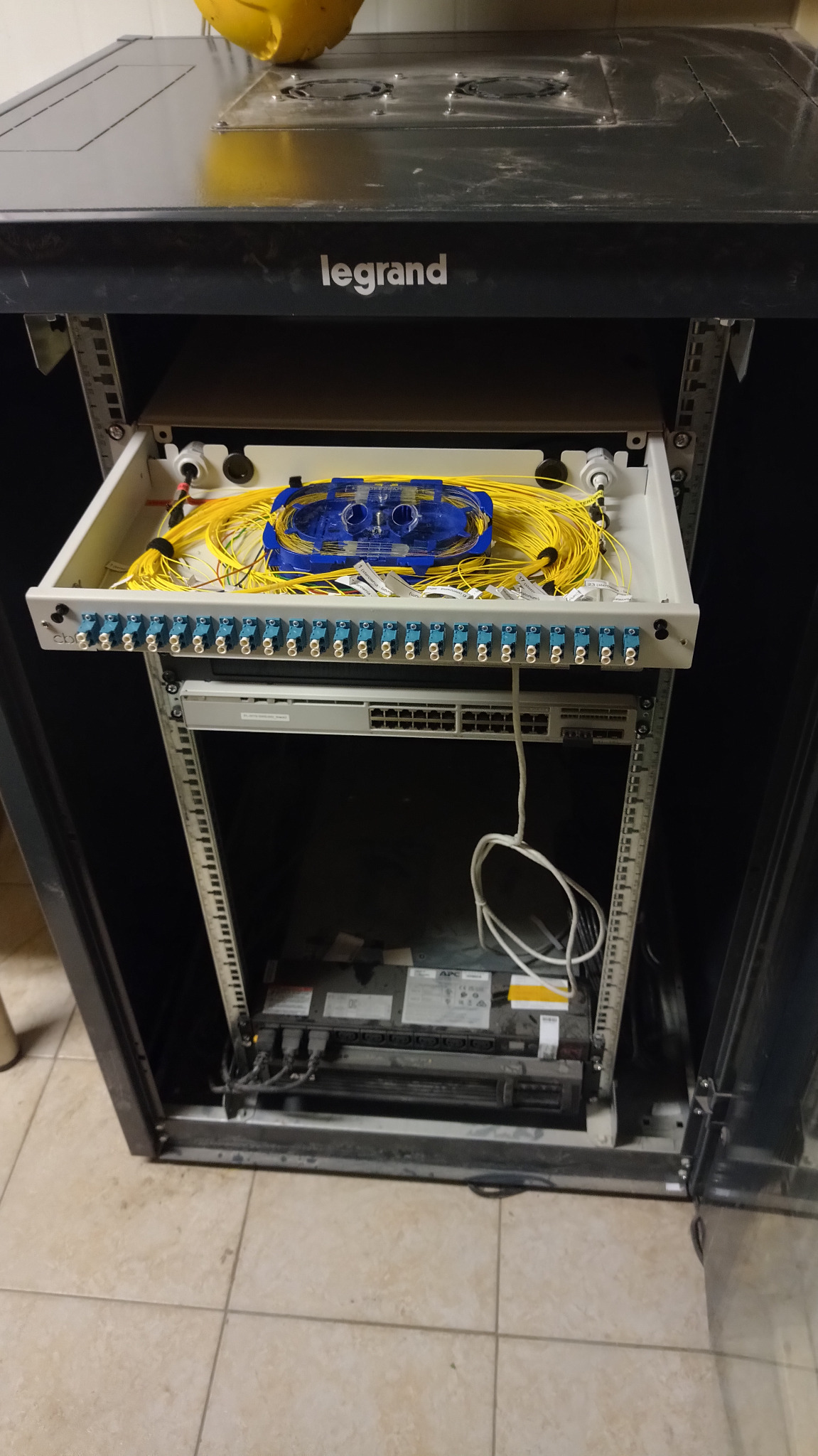 Szafa rack Legrand z wysuwaną półką zawierającą patch panel światłowodowy z żółtymi kablami światłowodowymi i niebieską kasetą spawów.