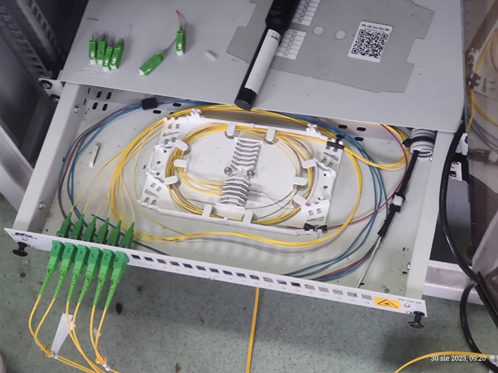 Rozdzielnica światłowodowa z uporządkowanymi kablami światłowodowymi wewnątrz, widoczne złącza SC/APC w kolorze zielonym i narzędzie do czyszczenia światłowodów na górze.