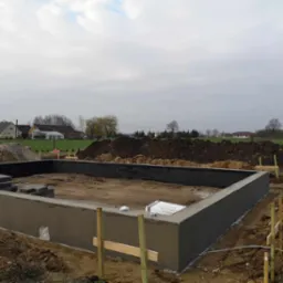 Fundamenty domu w trakcie budowy: wylany betonowy fundament z częściowo wymurowanymi ścianami z bloczków, widoczny stos bloczków oraz worki z materiałami budowlanymi, ziemia wokół wykopu...