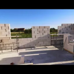 Wznoszone ściany domu z białych bloczków betonowych, widoczne rusztowania pomocnicze i zapas bloczków, w tle zieleń i zabudowania.
