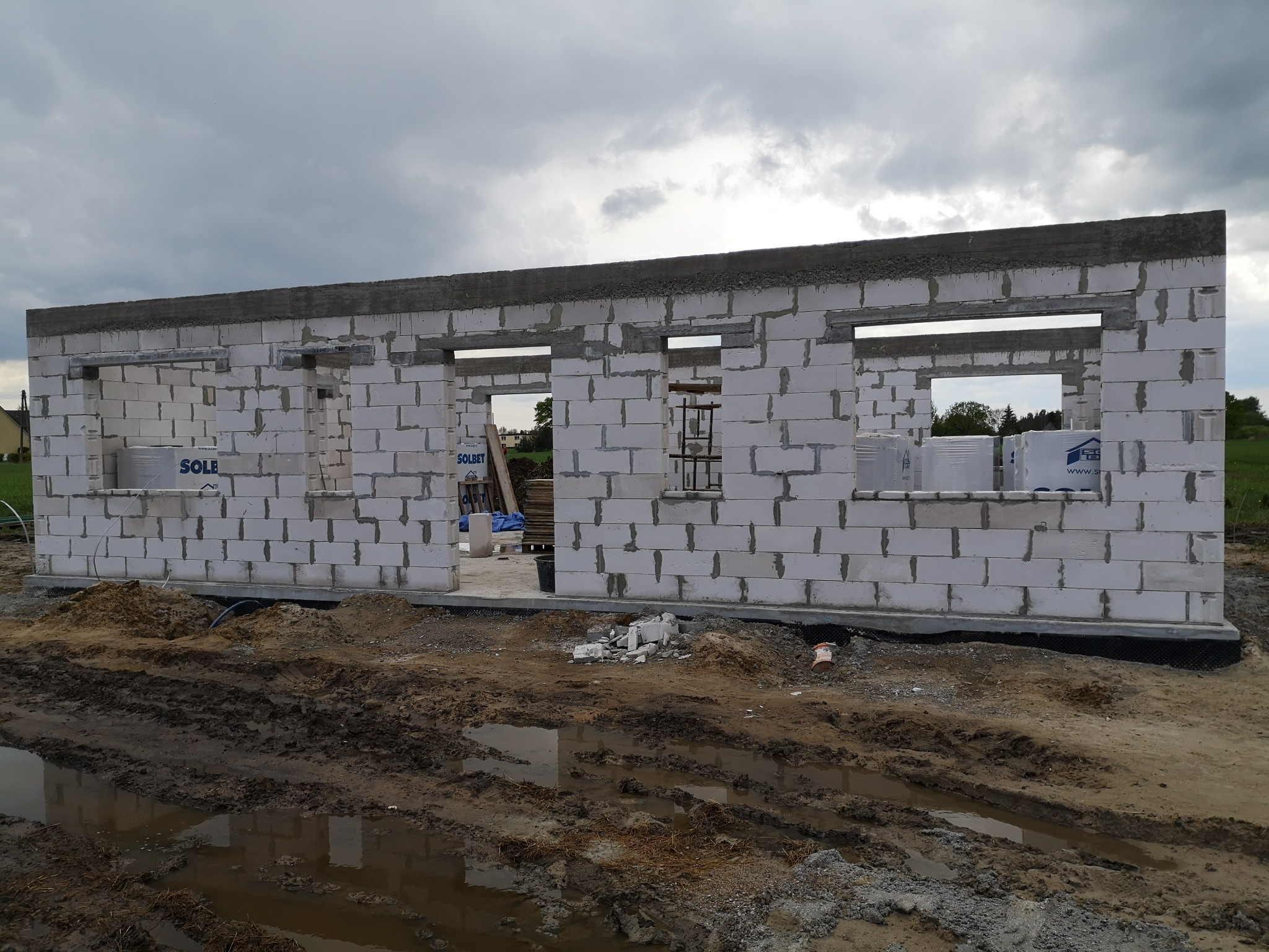 Wzniesione ściany parterowego budynku z betonu komórkowego z widocznymi otworami okiennymi i drzwiowymi, na fundamencie, z zapasami bloczków Solbet na placu budowy, w tle pochmurne niebo.