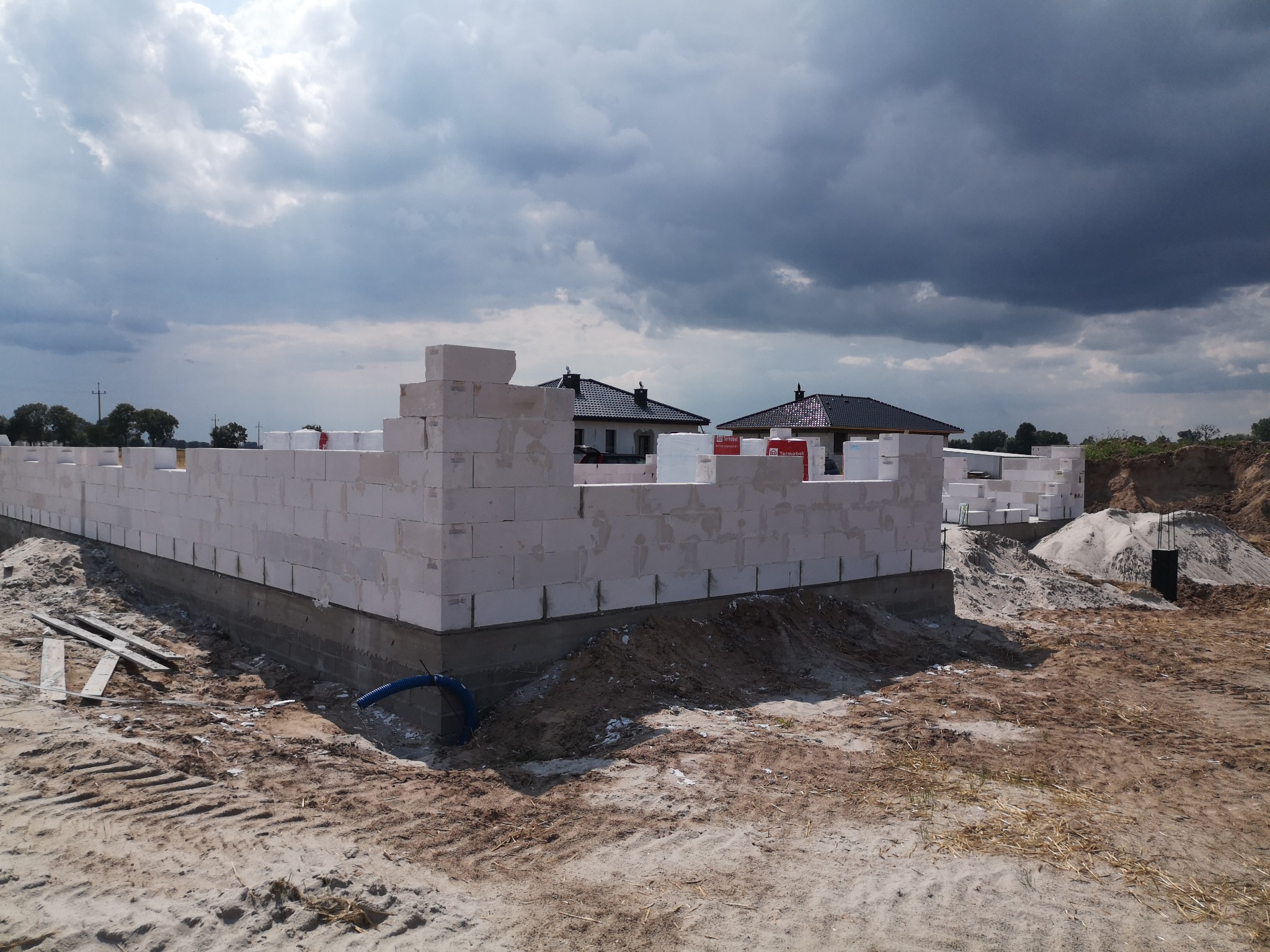 Wznoszenie ścian parterowego domu z białych bloczków na fundamencie, widoczny niebieski wąż przy fundamencie, w tle dwa ukończone domy jednorodzinne i pochmurne niebo.