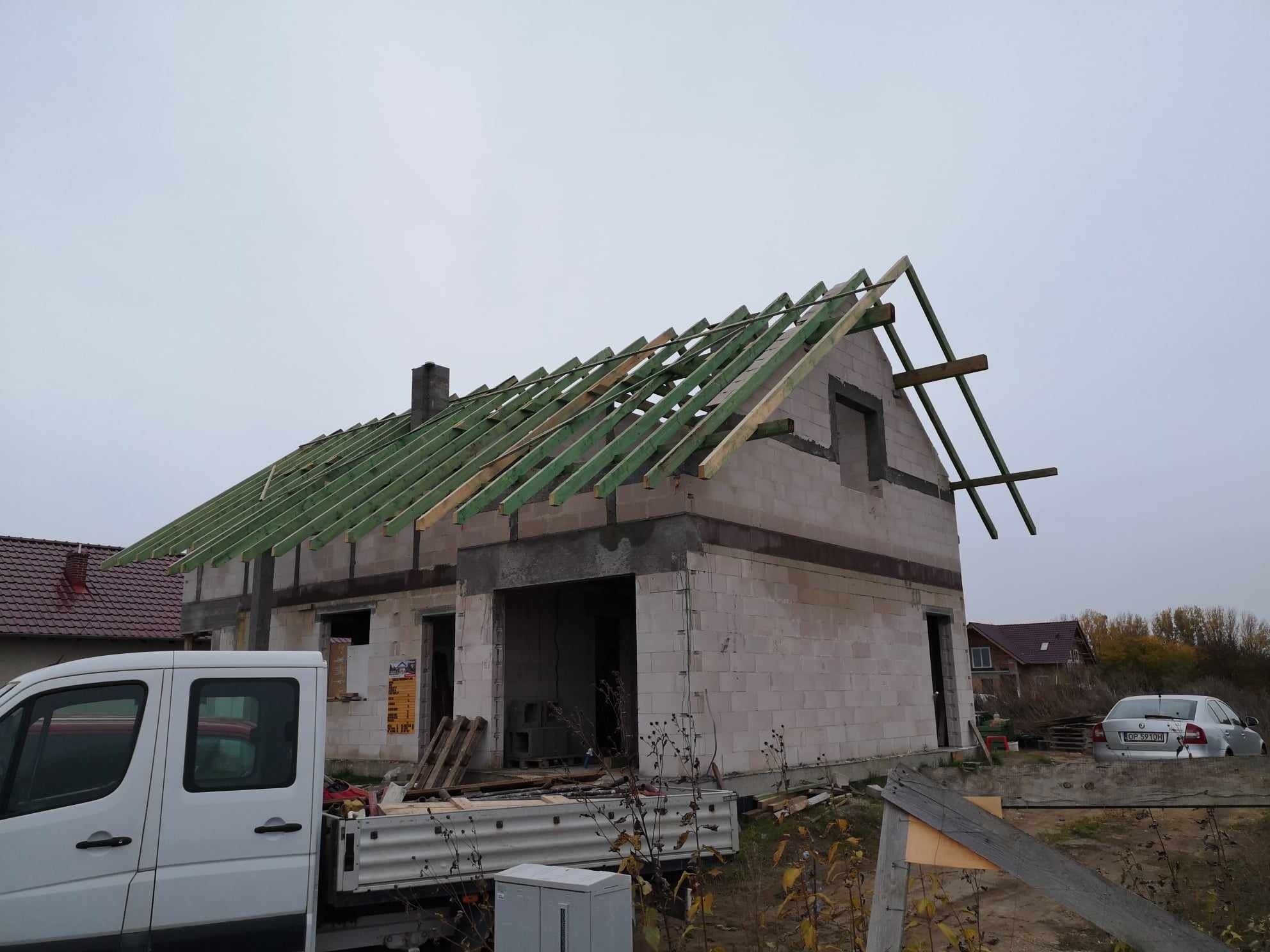 Surowy stan budynku z białych bloczków, z częściowo wykonaną konstrukcją dachu z zielonych łat, widoczny samochód dostawczy i materiały budowlane.