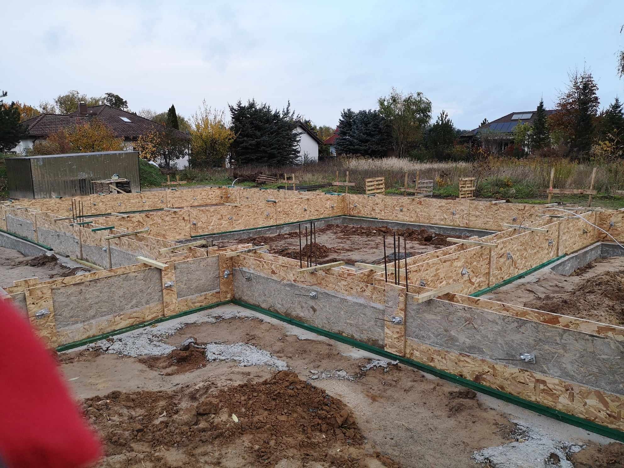 Wykop pod fundamenty z deskowaniem z płyty OSB, widoczne zbrojenie pionowe i zielona izolacja pozioma, w tle zabudowa jednorodzinna i pochmurne niebo.