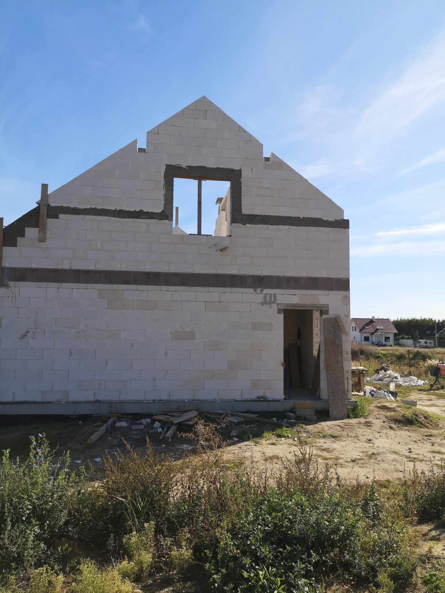 Surowy stan budynku jednorodzinnego z betonu komórkowego, widoczne zbrojenie nadproży, w tle inne budynki w oddali.