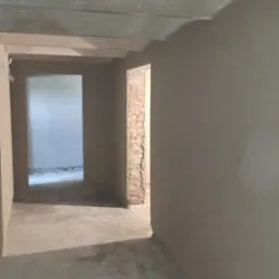 Wnętrze w trakcie prac wykończeniowych w Poznaniu: ściany pokryte świeżym tynkiem, widoczne surowe cegły w otworze drzwiowym, szare podłoże, sufit z elementami konstrukcyjnymi.