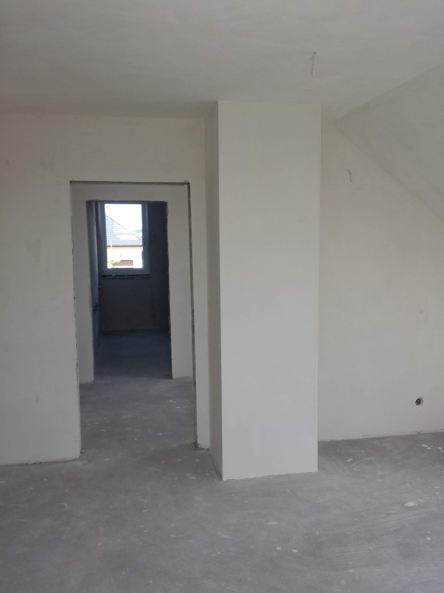 Wnętrze w stanie deweloperskim z surowymi ścianami i podłogą, widoczne przygotowane ościeże drzwiowe i fragment okna w tle.