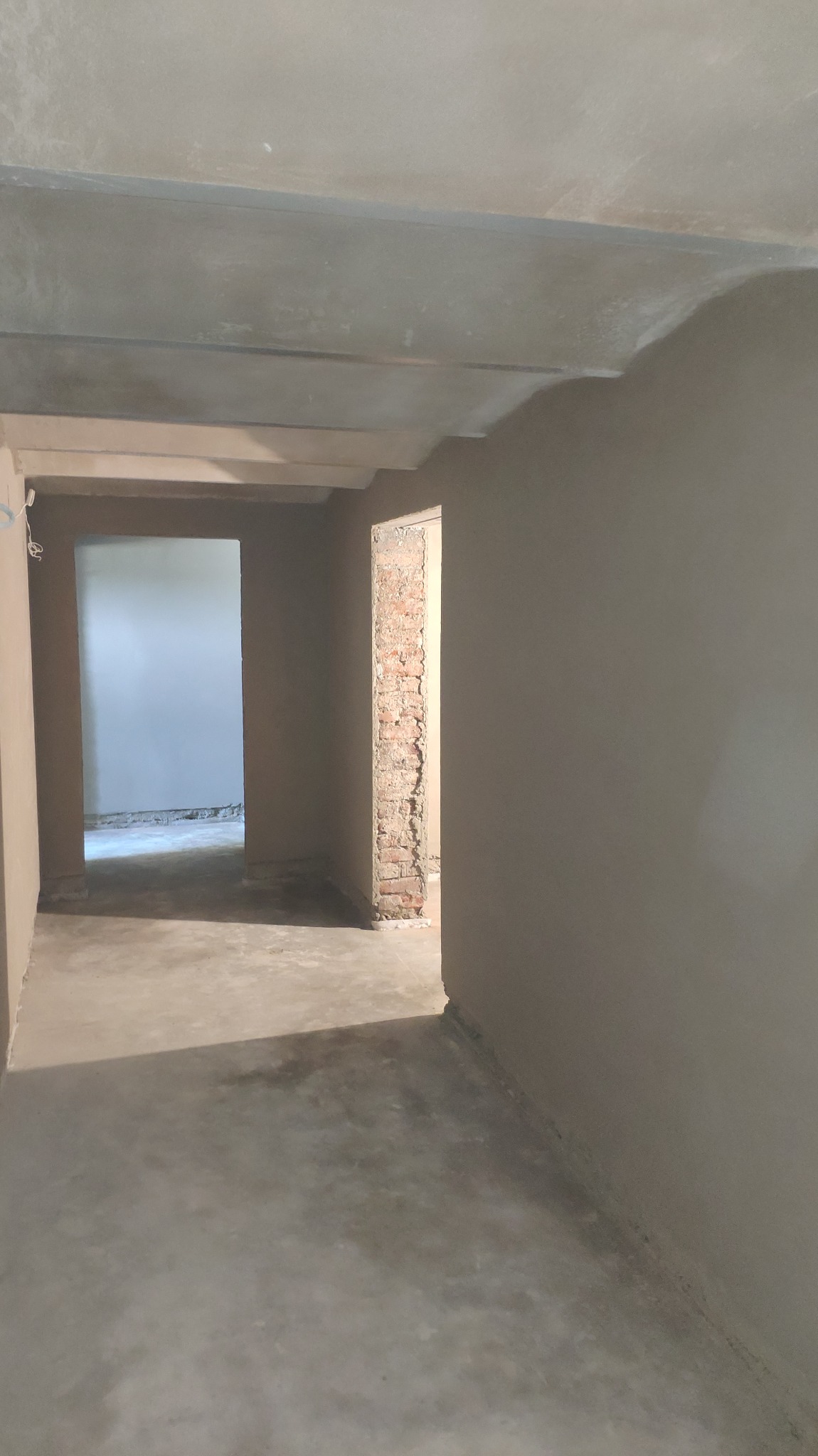 Wnętrze w trakcie prac wykończeniowych w Poznaniu: ściany pokryte świeżym tynkiem, widoczne surowe cegły w otworze drzwiowym, szare podłoże, sufit z elementami konstrukcyjnymi.