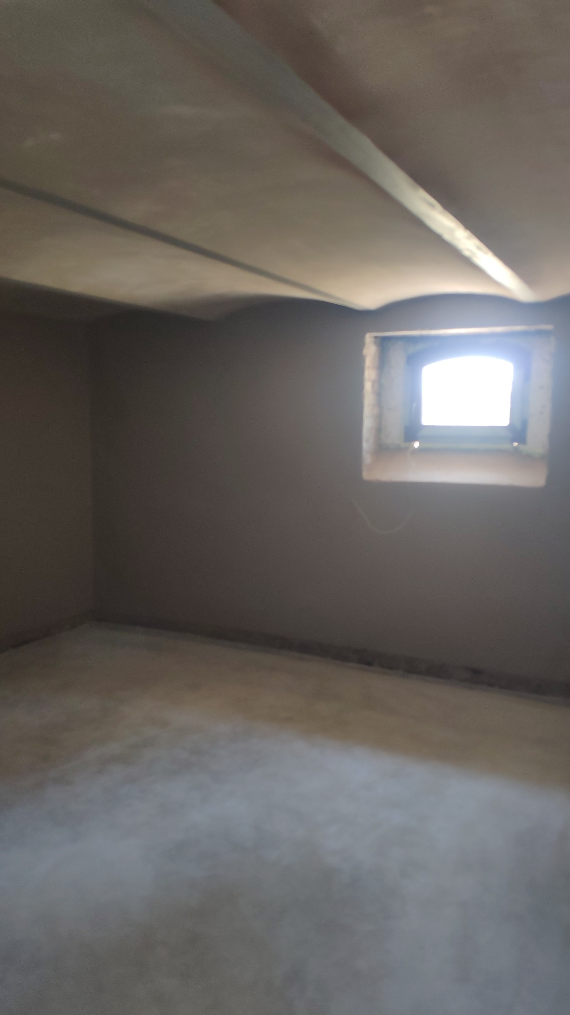 Pokój po tynkowaniu maszynowym, widoczne gładkie ściany, sufit z profilem, okno piwniczne z widocznym światłem dziennym.