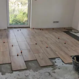 Układanie podłogi z płytek imitujących drewno w jasnym pomieszczeniu, widoczne poziomice i system poziomowania płytek z pomarańczowymi klinami, wiadro z zaprawą i skrzynka z narzędziami w rogu.