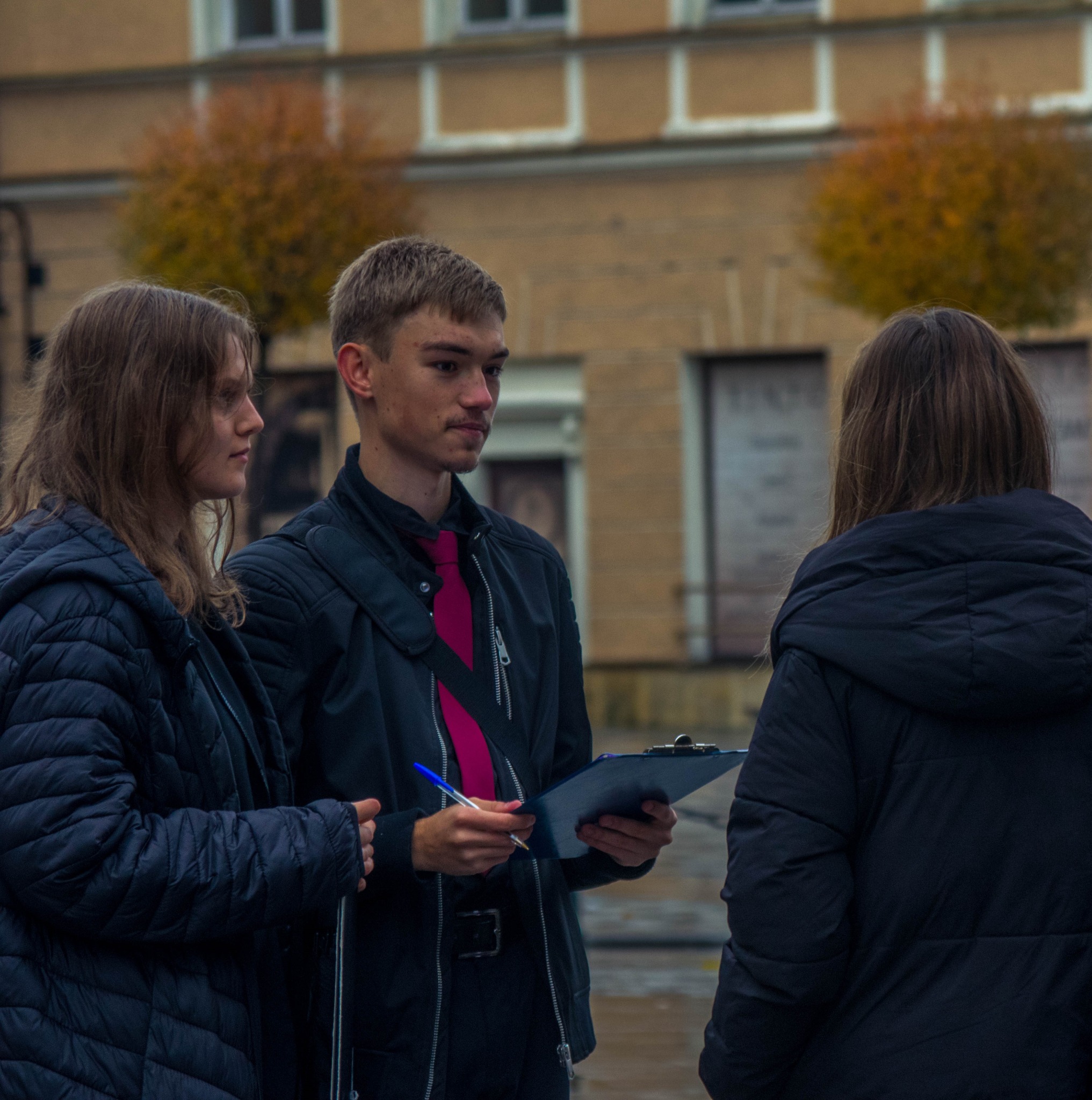 Młody mężczyzna w czarnej kurtce z różowym krawatem, trzymający niebieski długopis i podkładkę z dokumentami, rozmawia z dwiema kobietami na tle miejskiego budynku z jesiennymi drzewami.