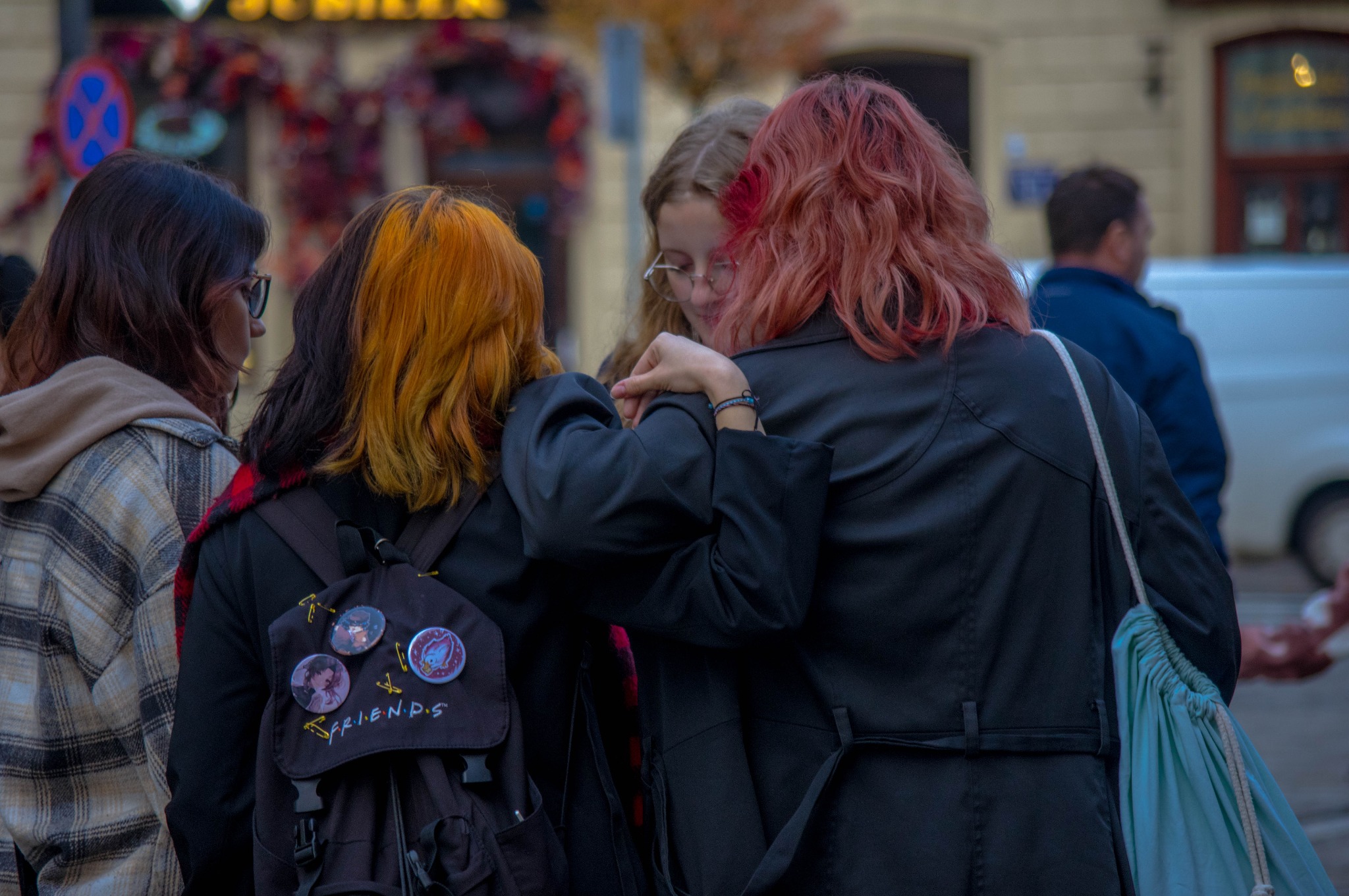 Grupa czterech młodych kobiet w miejskiej scenerii, z których trzy stoją blisko siebie, z widocznymi fryzurami w odcieniach brązu, blondu i czerwieni; jedna z nich ma plecak z przypinkami i napisem...