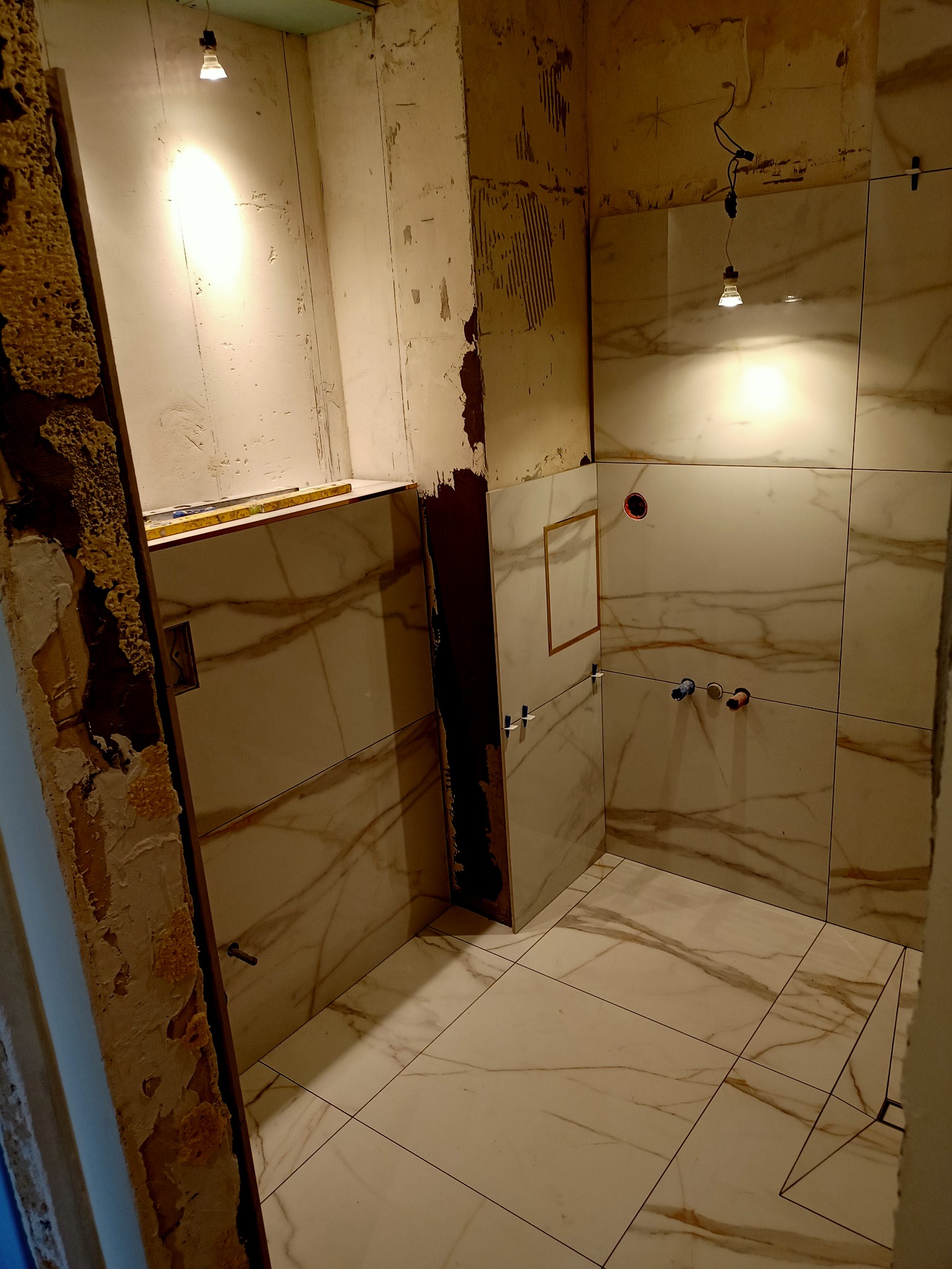 Łazienka w trakcie remontu: ułożone marmurowe płytki na ścianach i podłodze, widoczne instalacje, surowa ściana z resztkami starej okładziny, prowizoryczne oświetlenie.
