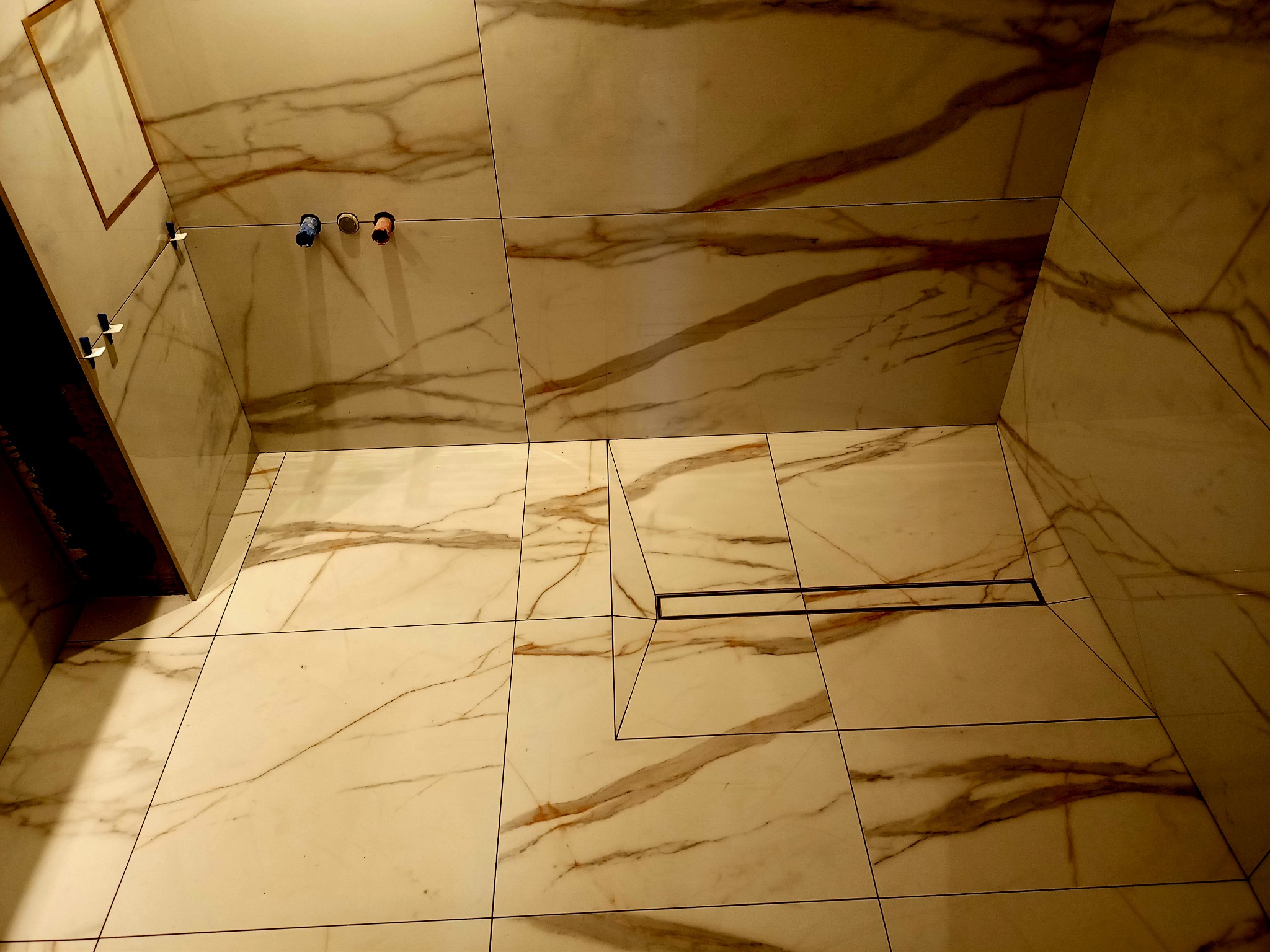Ułożone płytki imitujące marmur na podłodze i ścianach łazienki, widoczny odpływ liniowy w podłodze, krzyżyki dystansowe i wystające rury instalacji wodnej.