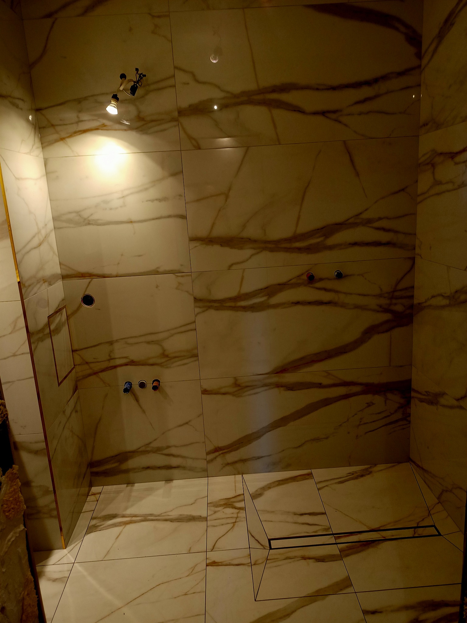 Wnętrze kabiny prysznicowej z marmurowymi płytkami, widoczne otwory na armaturę i odpływ liniowy w podłodze, oświetlenie punktowe na suficie.