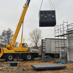 Kma Energy - Żółty dźwig podnosi rozdzielnicę elektryczną na placu budowy, widoczne rusztowania i podłoże z piasku.