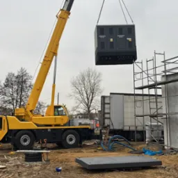 Żółty dźwig podnosi rozdzielnicę elektryczną na placu budowy, widoczne rusztowania i podłoże z piasku.