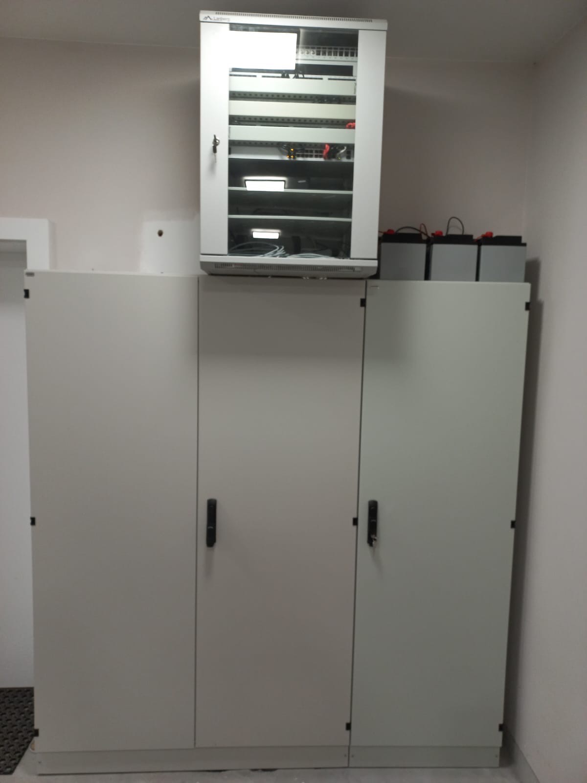 Szafa serwerowa z przeszklonymi drzwiami, wypełniona sprzętem sieciowym, umieszczona na szafkach z trzema parami drzwi, zasilana trzema akumulatorami na górze, w pomieszczeniu technicznym.