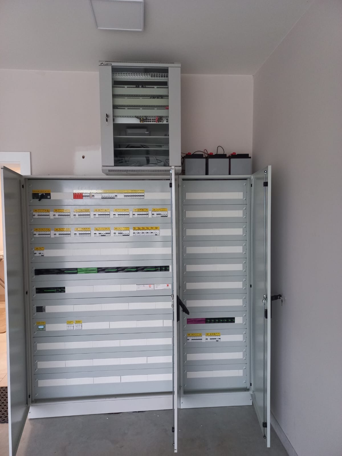 Otwarta, duża szafa rozdzielcza elektryczna z widocznymi bezpiecznikami i okablowaniem, umieszczona w jasnym pomieszczeniu, z dodatkową mniejszą szafką powyżej i trzema akumulatorami na jej szczycie.