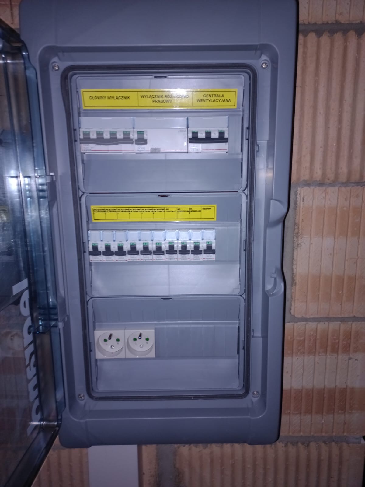 Otwarta szara skrzynka rozdzielcza z widocznymi bezpiecznikami, wyłącznikami i gniazdkami, zamontowana na ścianie z cegły.
