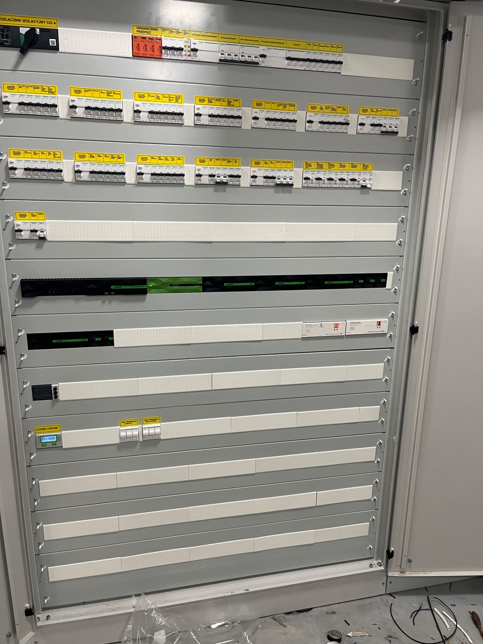 Rozdzielnica elektryczna z zamontowanymi zabezpieczeniami, wyłącznikami, ogranicznikiem przepięć i licznikiem Loxone, widok z bliska na szare panele i elementy instalacji elektrycznej.