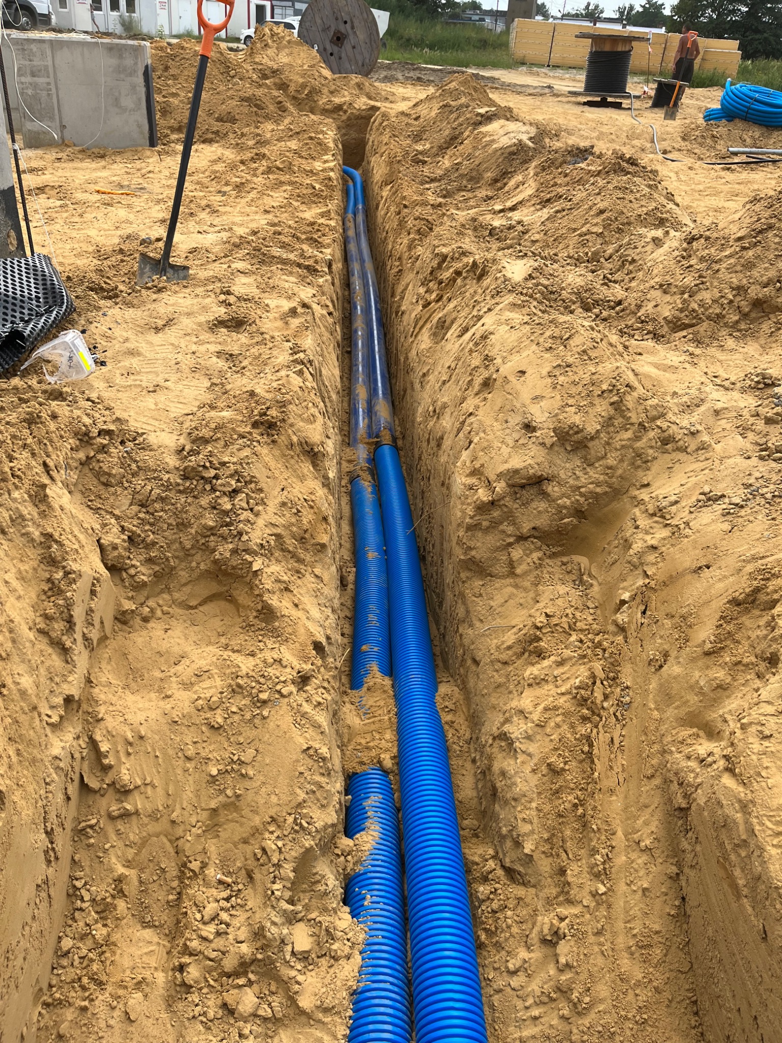 Głęboki wykop w ziemi, w którym ułożone są niebieskie rury karbowane, szpadel wbity w ziemię na skraju wykopu, w tle widoczny budynek i drewniana szpula kablowa.