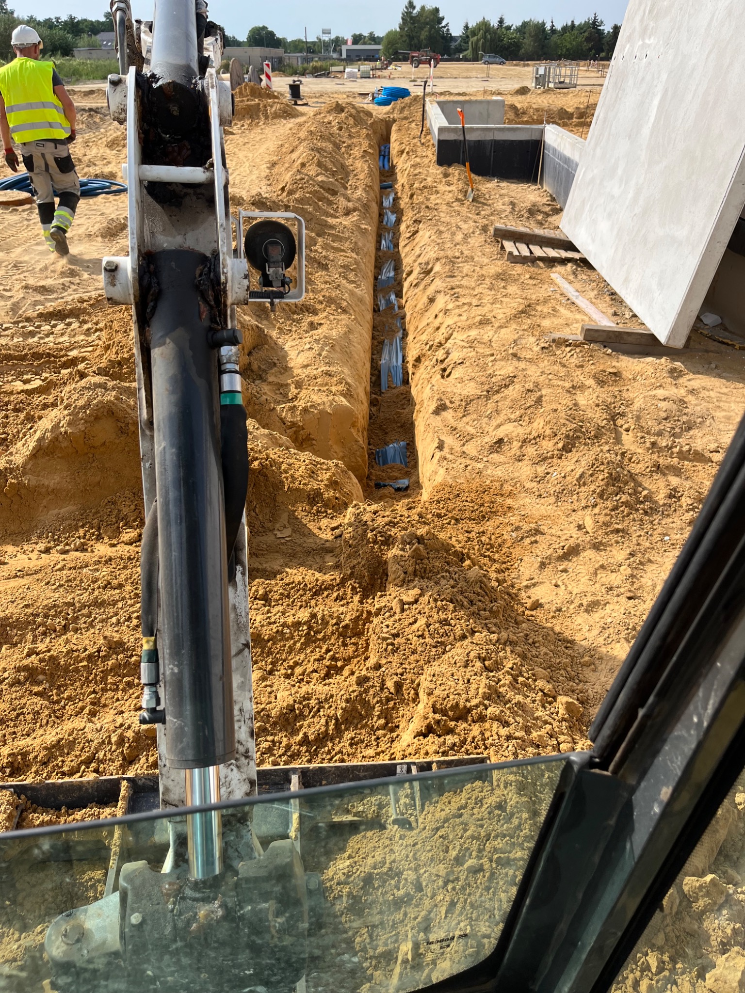 Wykopany rów z ułożonymi rurami instalacyjnymi, widok z kabiny koparki, w tle fundamenty budynku i pracownik w kamizelce odblaskowej.