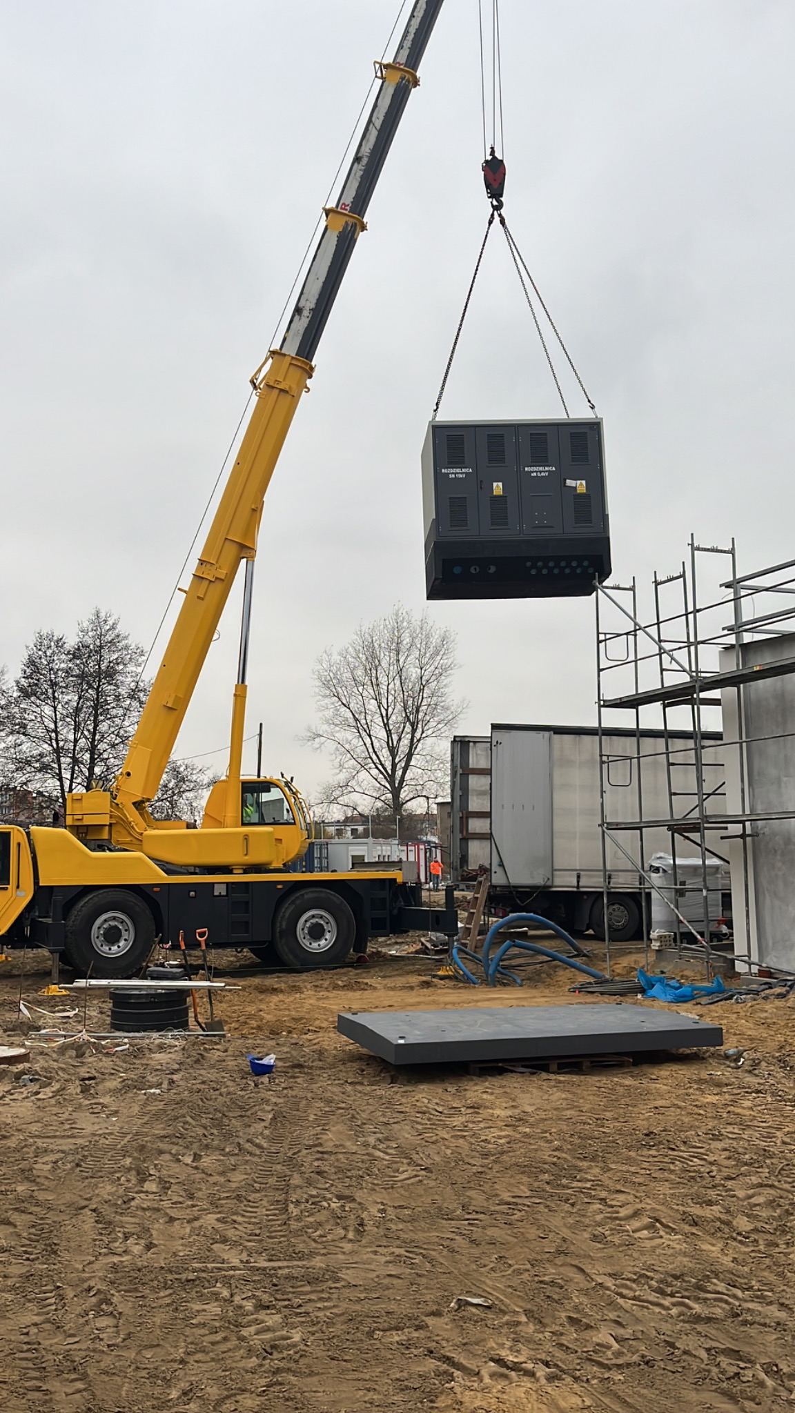 Żółty dźwig podnosi rozdzielnicę elektryczną na placu budowy, widoczne rusztowania i podłoże z piasku.
