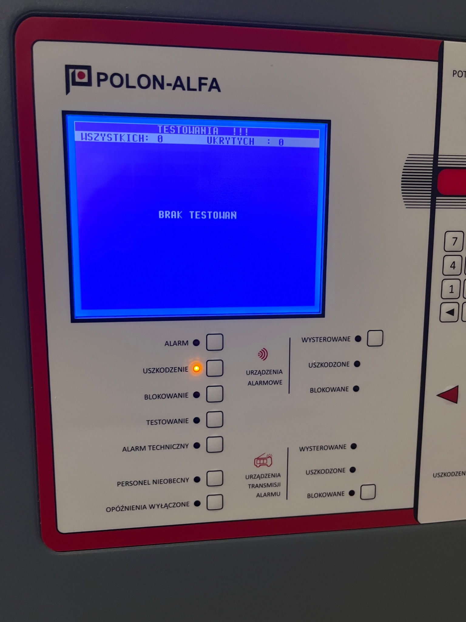 Panel sterowania systemem alarmowym Polon-Alfa z podświetlonym na pomarańczowo wskaźnikiem 'Uszkodzenie' i komunikatem 'Brak Testowań' na niebieskim ekranie.