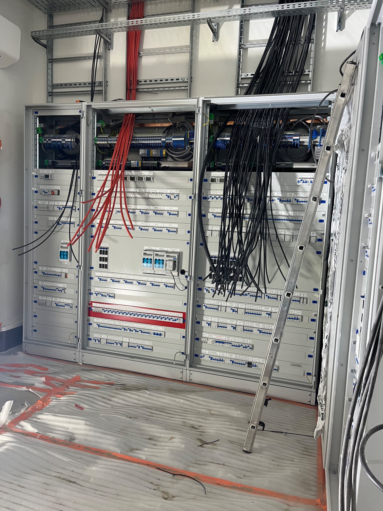 Rozdzielnica elektryczna w trakcie montażu, widoczne liczne przewody czerwone i czarne, drabina oparta o szafę, podłoga zabezpieczona folią malarską.